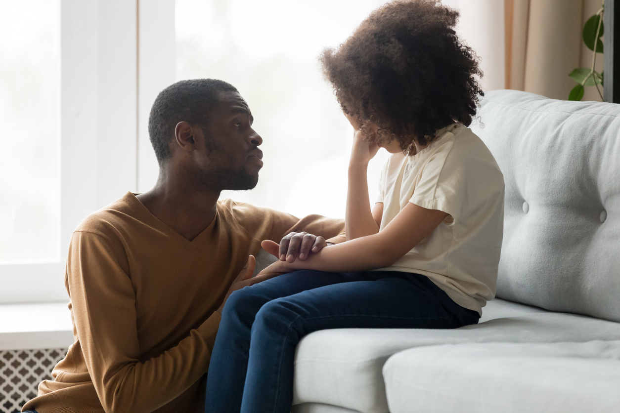 Child sitting on a couch looking sad and being comforted by dad sitting on floor