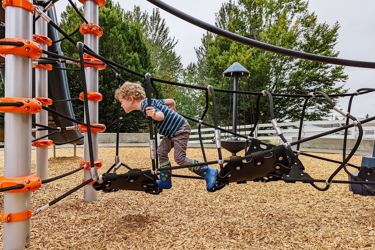 Young child runs through a rope tunnel toward a climbing tower at the new playground at Kent’s Chestnut Ridge Park