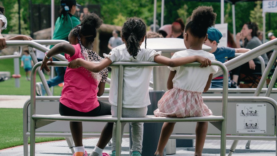 Three girls sit on the bench seat of an accessible ground-level merry-go-round at Seattle's Pathways Park