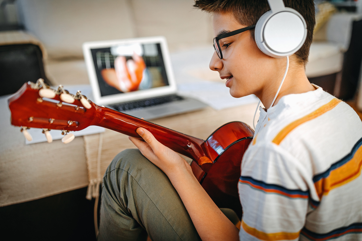 teenager sitting in front of a youtube video on a laptop holding a guitar