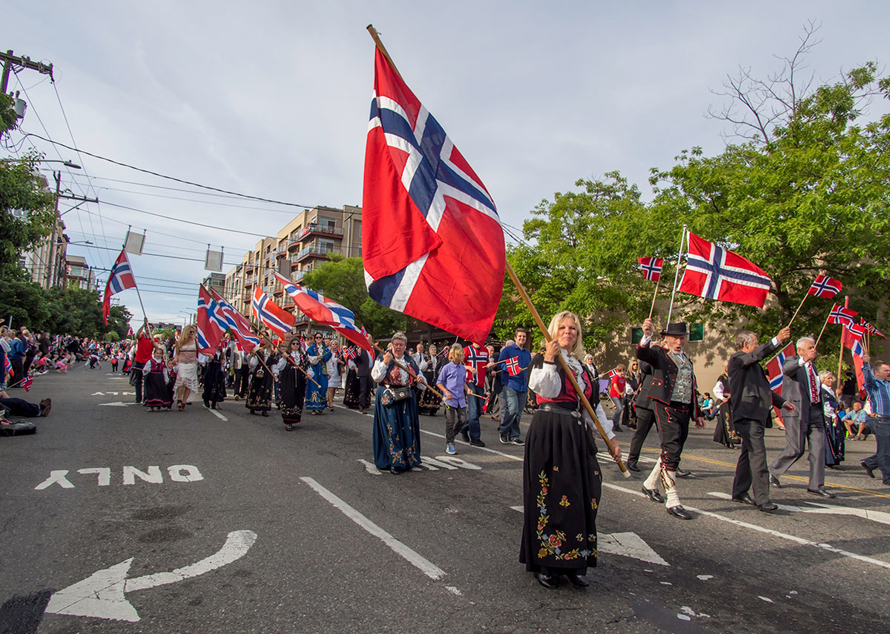 People in Norwegian traditional costumes walk in Ballard Syttende Mai parade holding Norwegian flags
