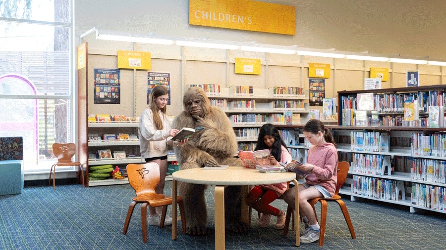 Kids at a library reading books with a person wearing a bigfoot costume