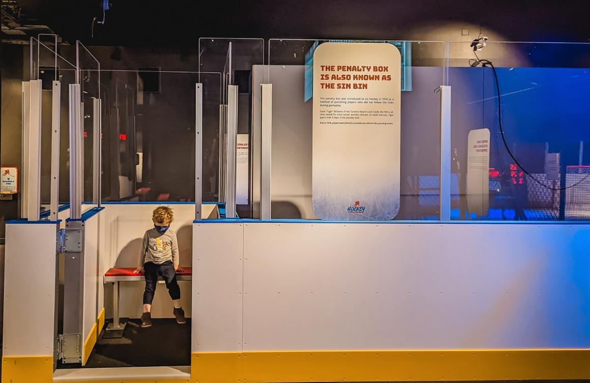A young boy of about 2 wears a mask and sits in model penalty box or sin bin at new exhibit Hockey: Faster Than Ever at Seattle's Pacific Science Center