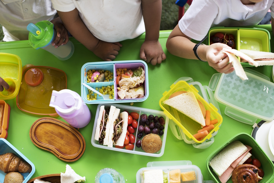 A view from above of a lunch table with lunches in a variety of lunch boxes at an elementary school.
