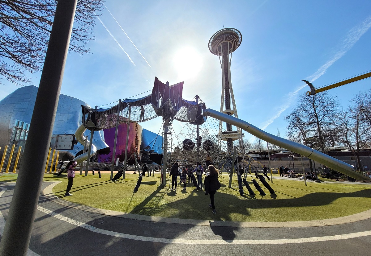 Kids play on the Artists at Play playground at Seattle Center on a sunny spring day. The Space Needle and the Museum of Pop Culture (MOPOP) are visible behind