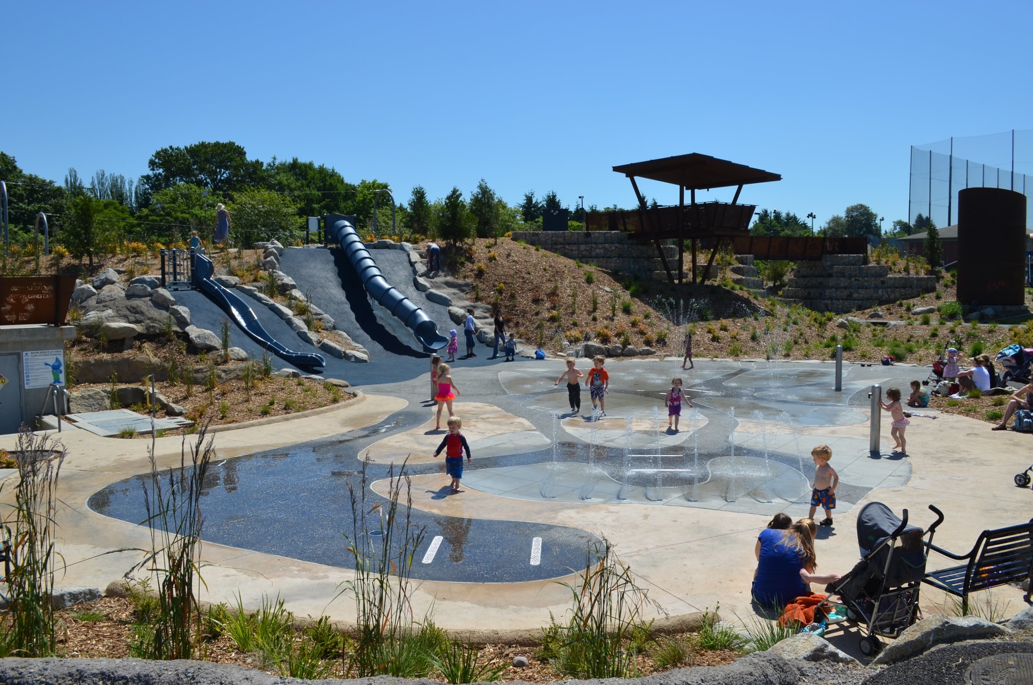 Kids play on the summer spray ground at Seattle’s Jefferson Park with two hillside slides in the background, Jefferson Park's playground is among Seattle's most adventurous and fun playgrounds
