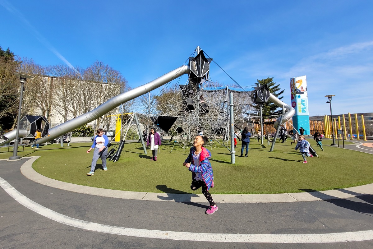 A girl runs in front of the high climber and long tube slide that are part of the Artists at Play playground at Seattle Center among risky adventure playgrounds for thrill-seeking kids