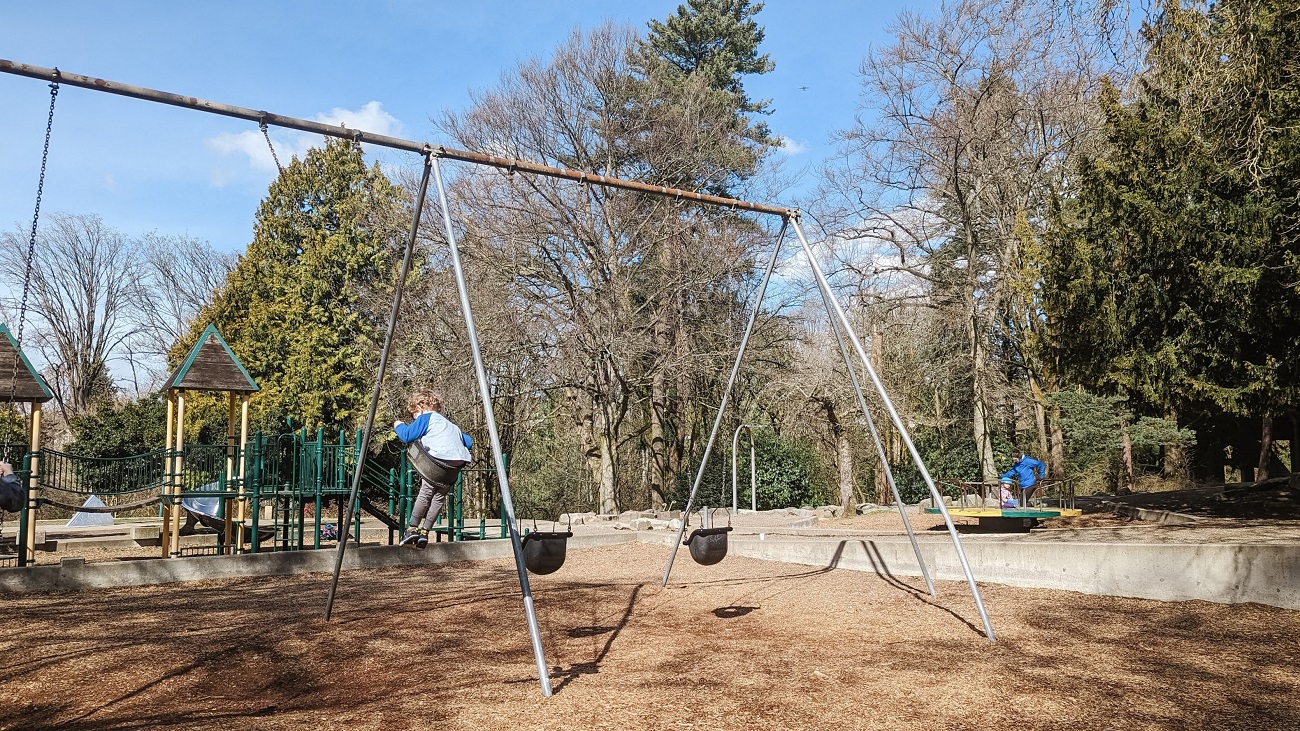 A boy swing high on longer swings at Seattle’s Cowen Park, with a castle-themed play structure, merry-go-round and zip line visible in the background