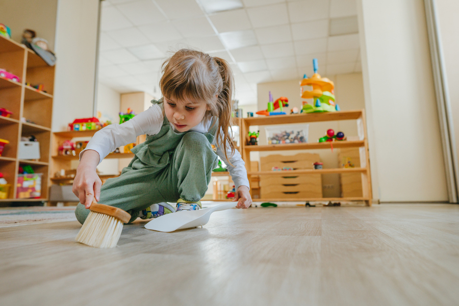 child sweeps up her Montessori playroom with organized low shelves in the background