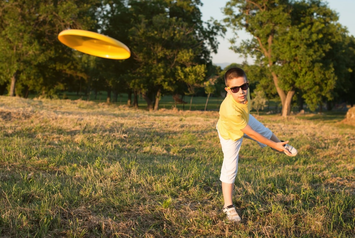 A school-age boy in a yellow shirt and sunglasses throws a yellow Frisbee disc during a game of disc golf free fun family sport to play in parks around Seattle