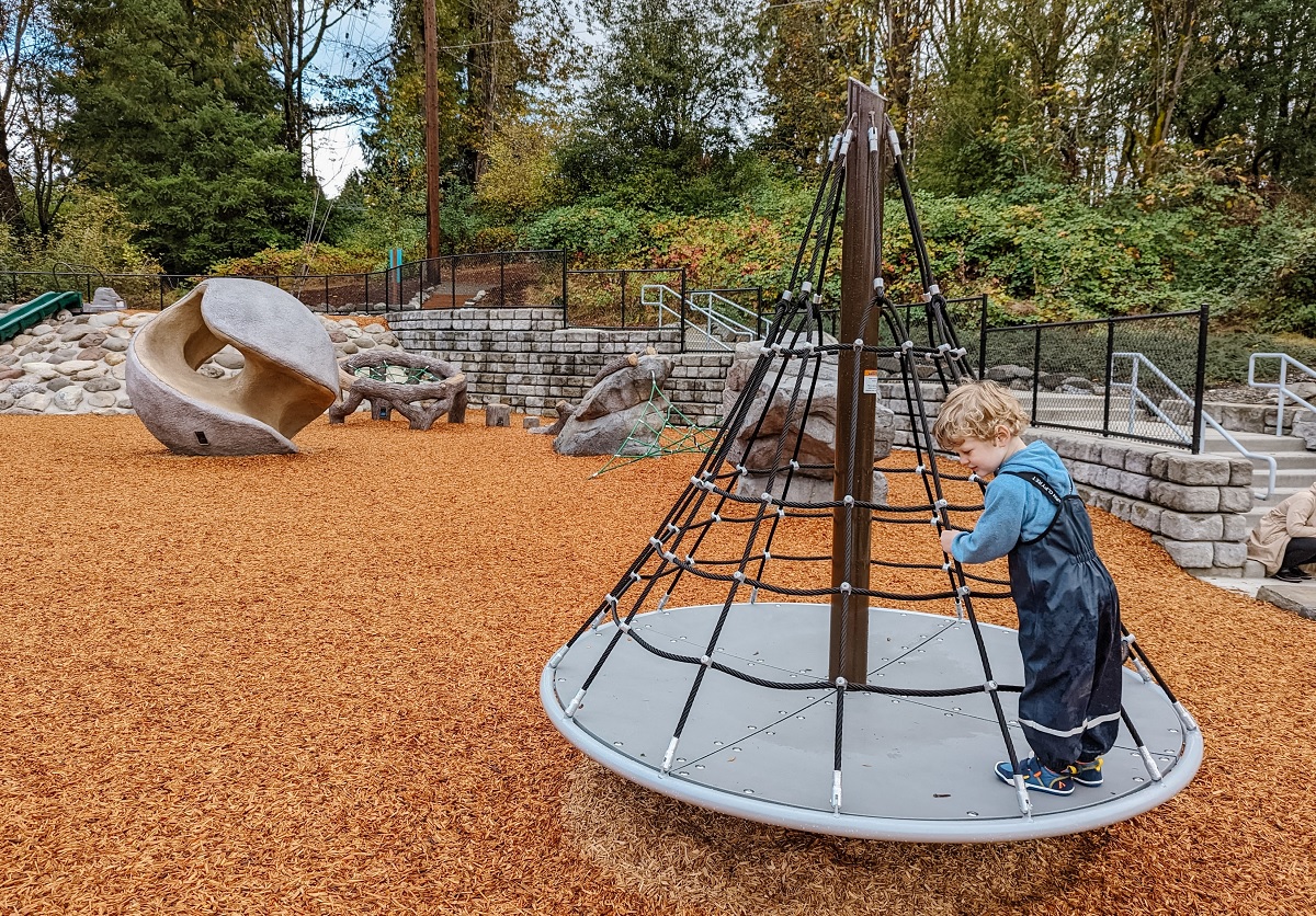 Young boy on cone shaped cargo net merry-go-round spinner at Newport Hills Woodlawn Park in Bellevue