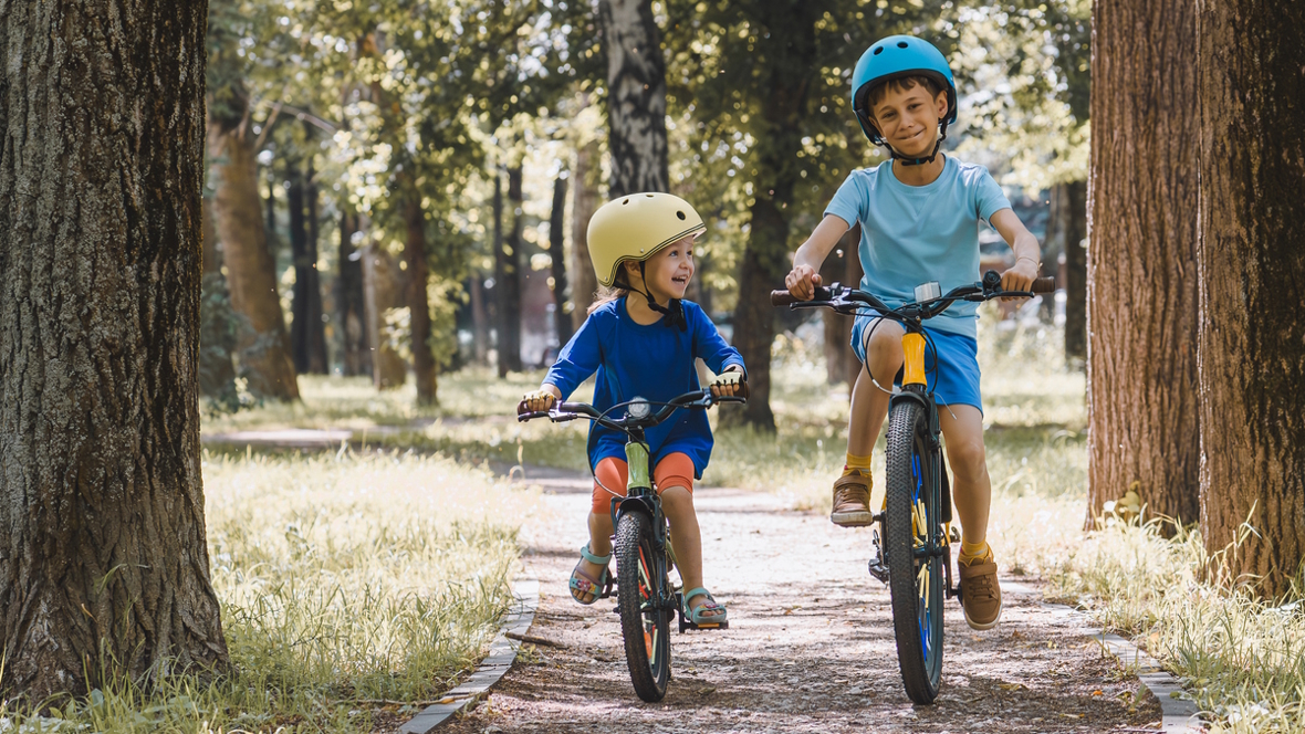 Two kids riding bikes on a path in the summer wearing helmets