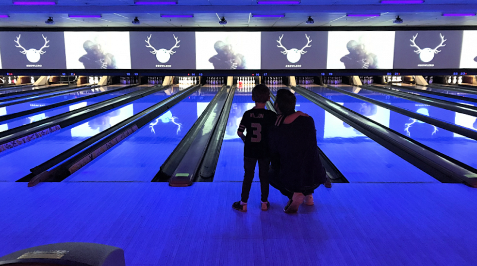 Crouching father alongside boy seen from the back. They're looking down a bowling lane in a bowling alley. The lighting is blue-ish.