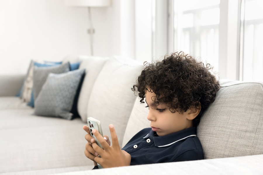 Young boy sitting on a couch alone looking at a smart phone might be part of the anxious generation