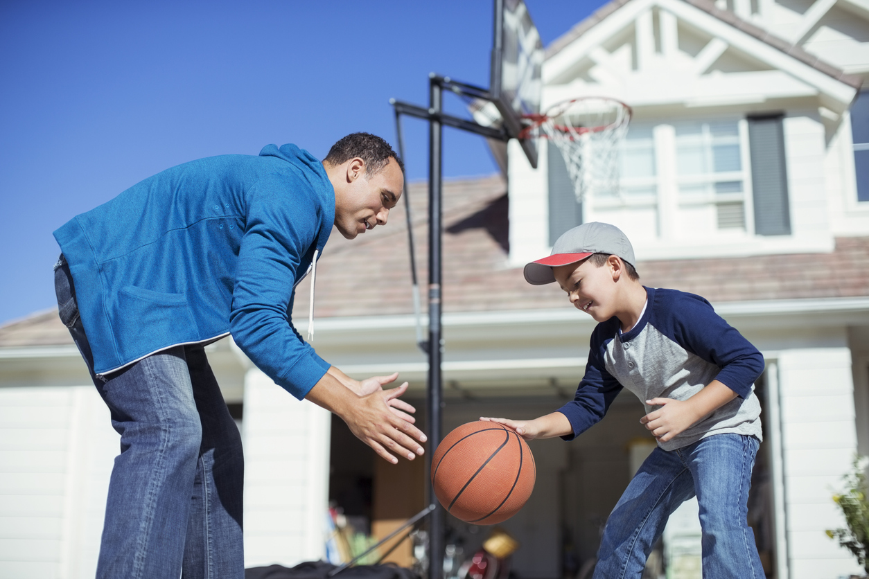 Father and son playing basketball outside of a big white house