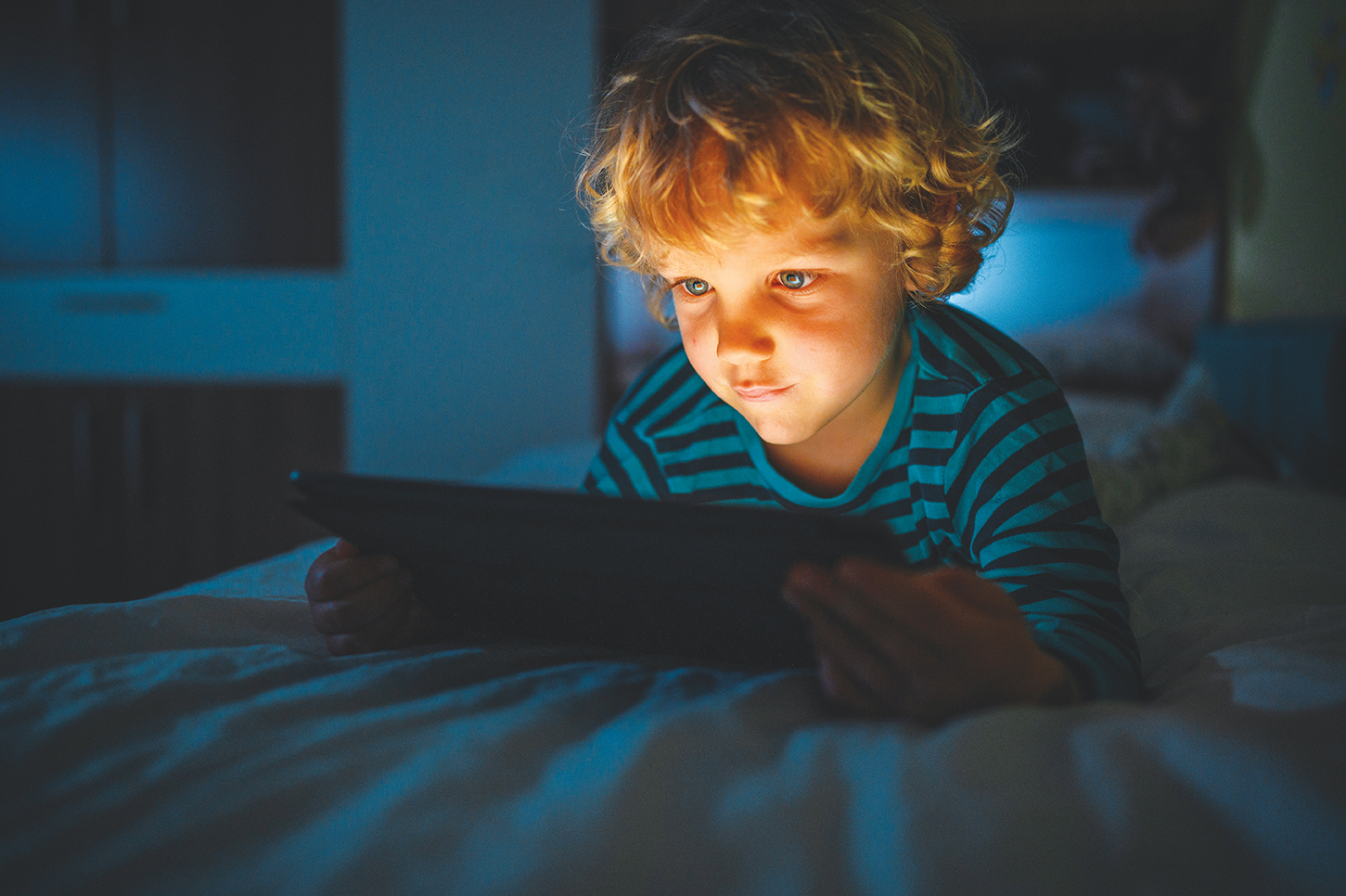 curly-haired blond boy laying on his bed in the dark with an ipad lighting up his face