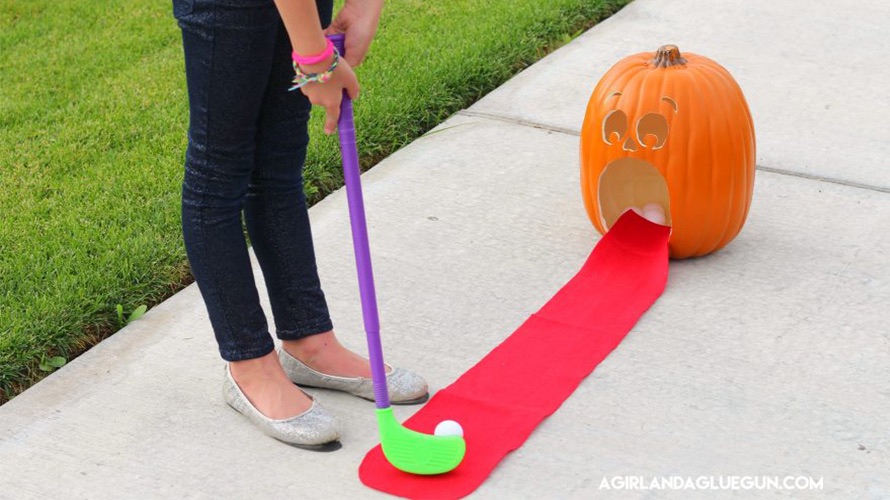 A kid putts a golf ball on a red carpet into a pumpkin mouth during a Hallowen party game