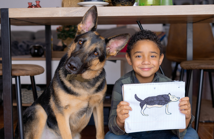 a boy sits next to his German Shepard proudly holding his drawing of the dog