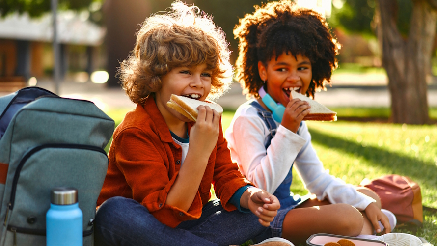 Two kids eating lunch together outside on a sunny day at school