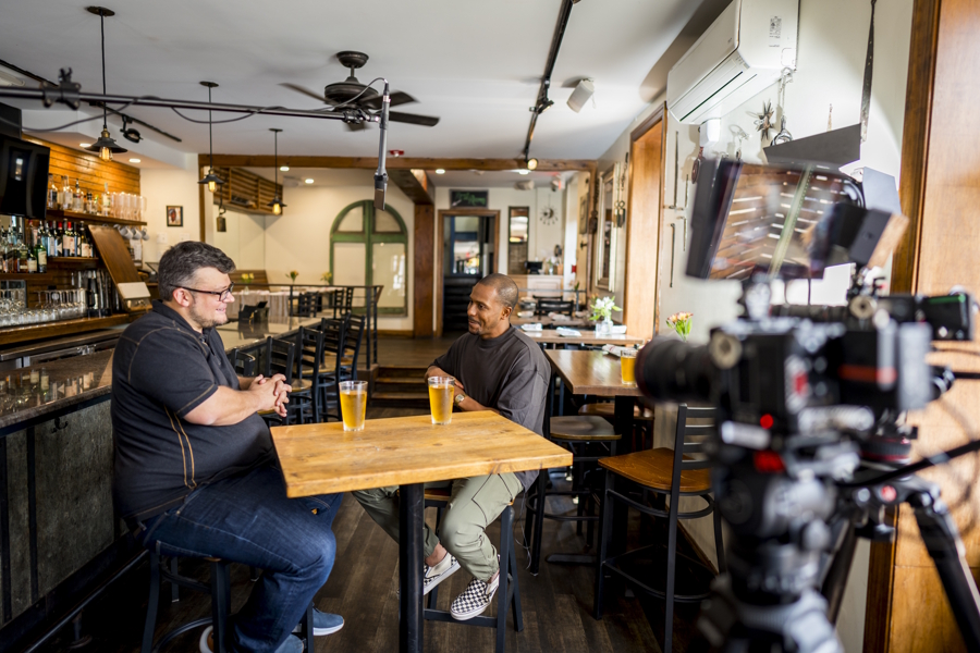 Two men sitting at a table talking and being filmed for a TV series