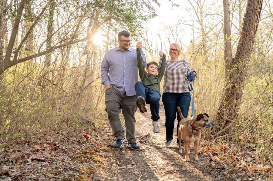 parents swinging their child while on a walk in the woods with a dog