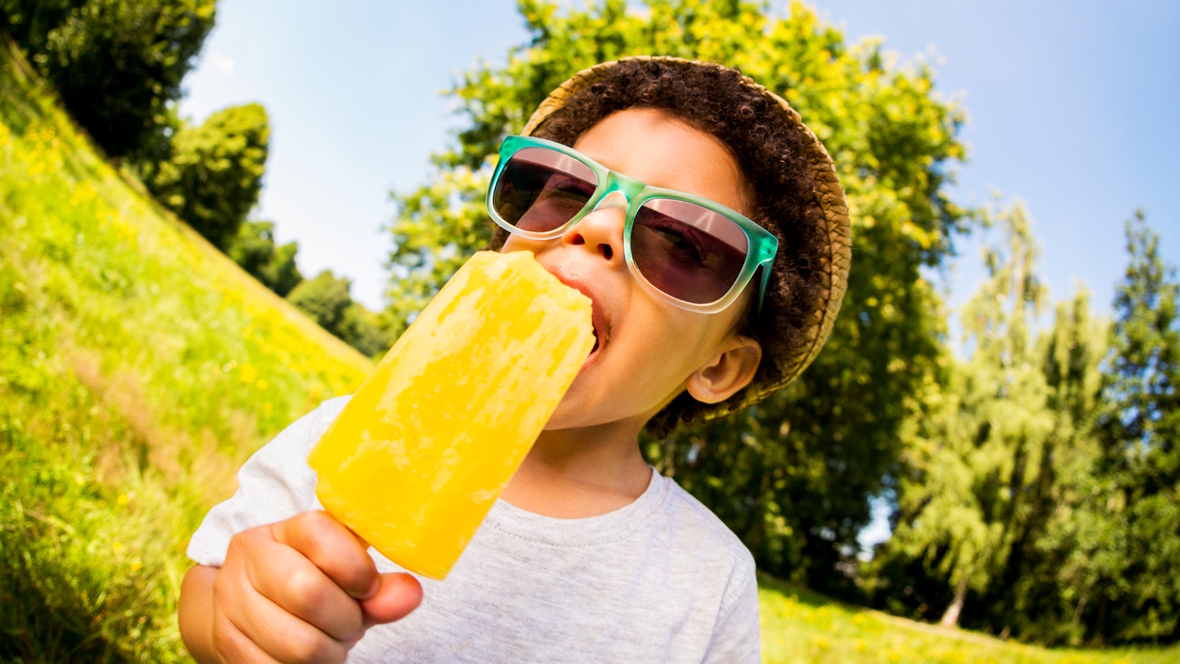 Boy wearing a hat and sunglasses eating a sweet treat in the summer