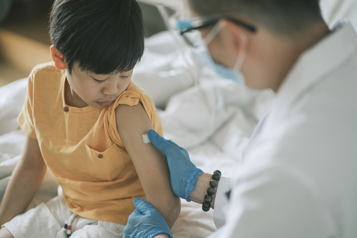 small boy looking down at his arm as a masked doctor puts a bandaid over a vaccine shot poke