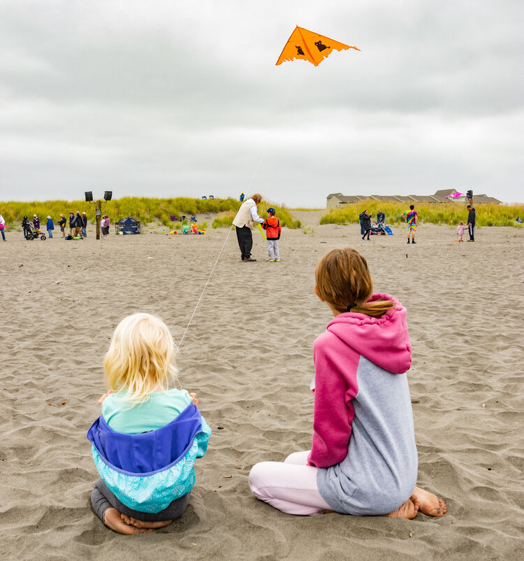 Two kids sitting in the sand flying a kite at the beach
