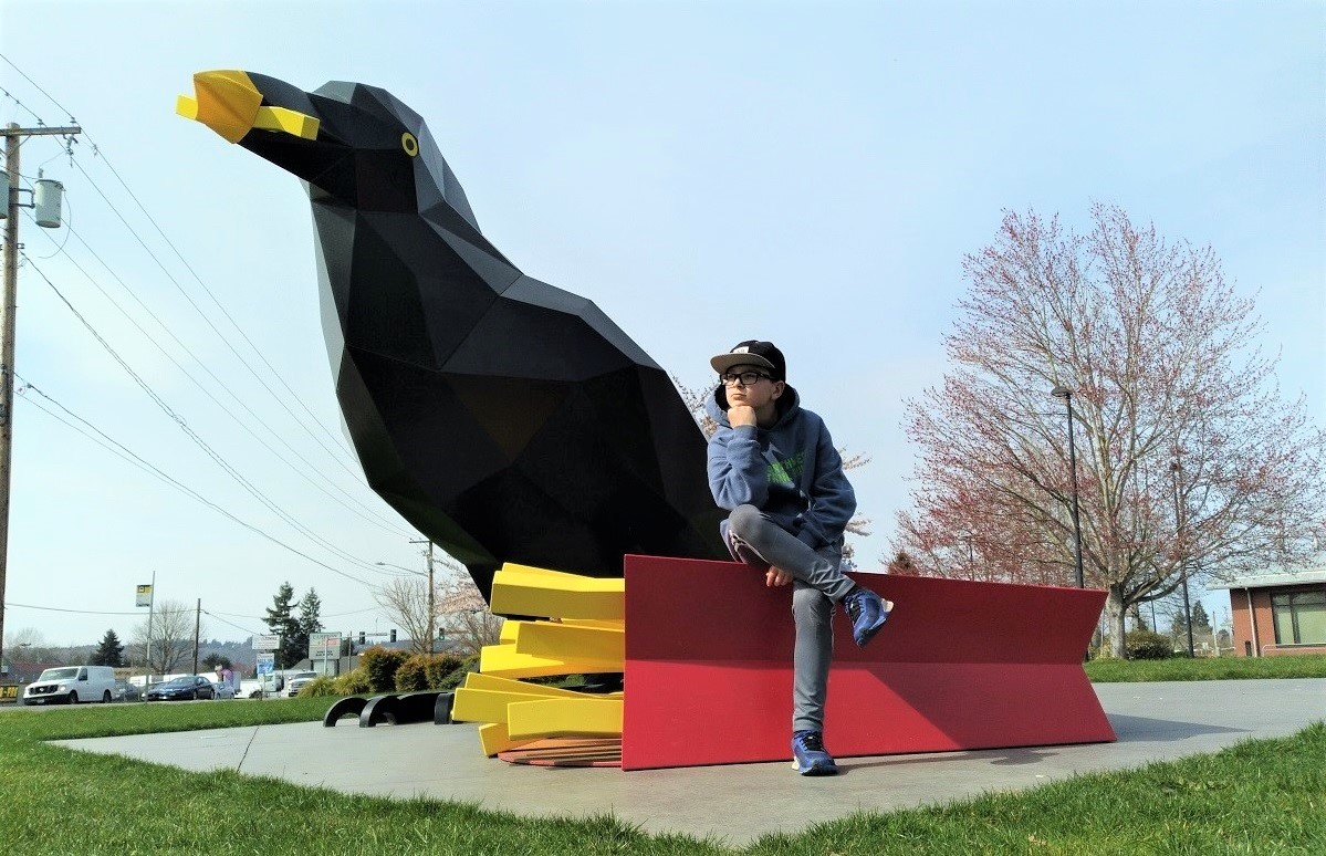 Young teenage boy in hat glasses sweatshirt seated on french fries of Auburn's Crow With Fries sculpture at Les Gove Park best instagram worth sites around Seattle