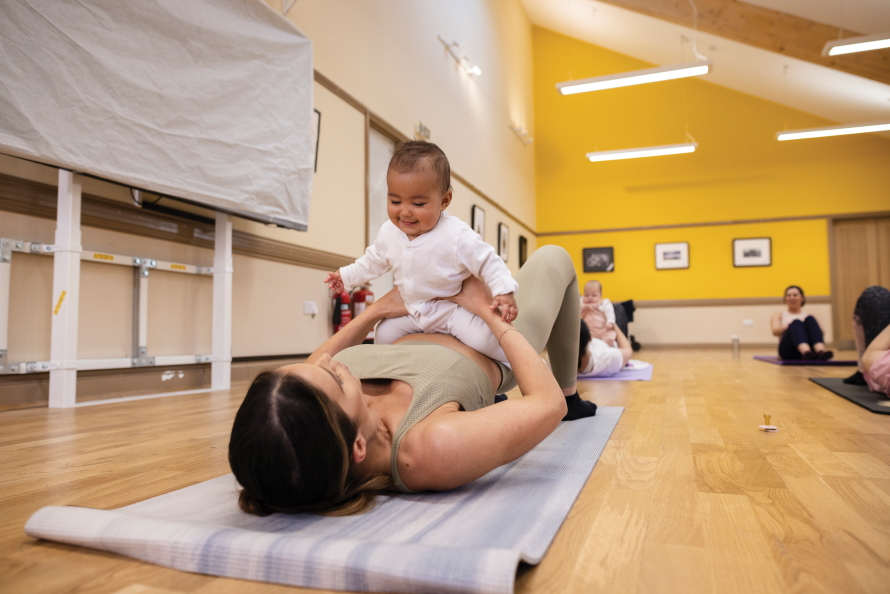 Woman laying on her back on a yoga mat with a baby smiling and sitting on her stomach