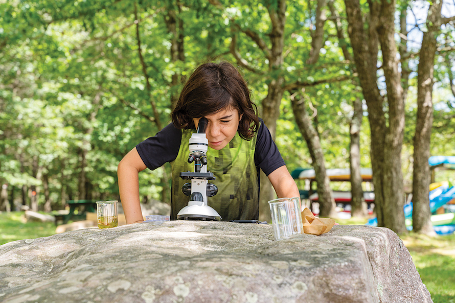 boy bent over a microscope resting on a large rock outside