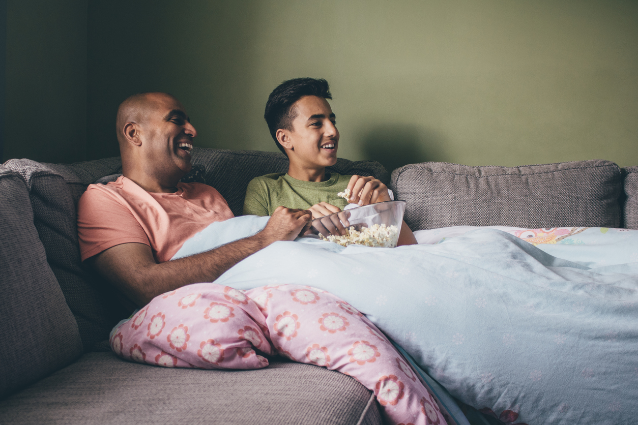 father and teen son under a blanket on the couch watching a movie