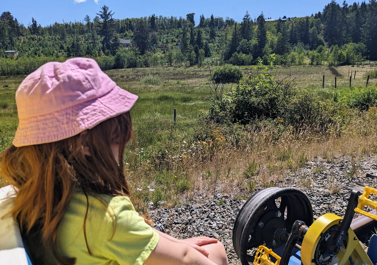 A girl who can’t reach the rail cycle pedals even with a booster rides along and enjoys the excursion