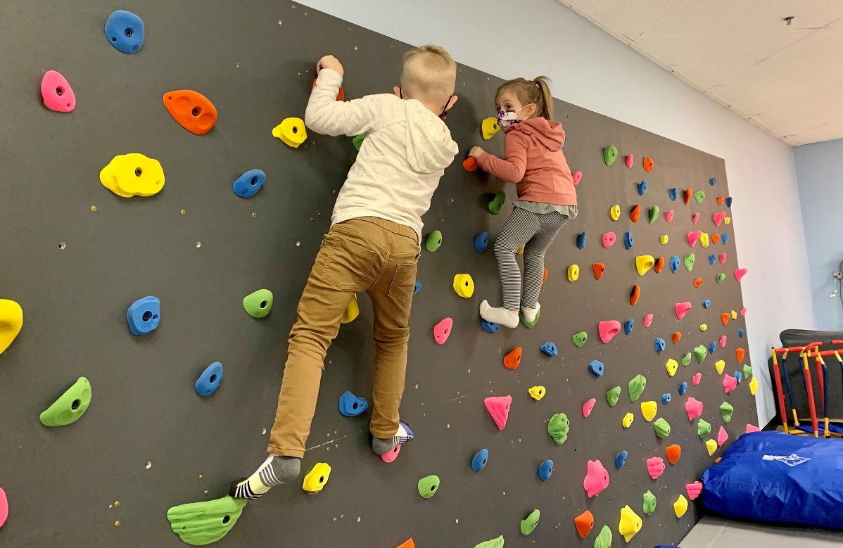 Two young kids wearing masks climb on an indoor climbing wall at Kaleidoscope Family Gym indoor play gym in Puyallup, Wash.
