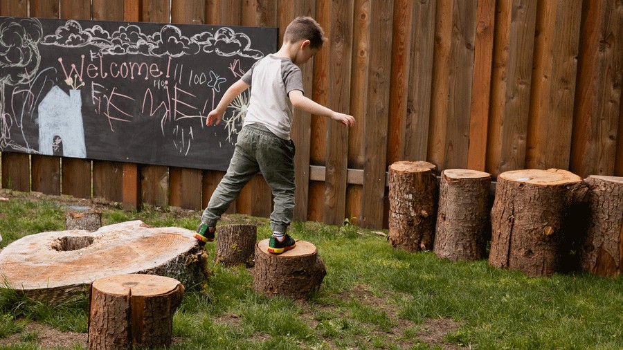 young boy walking across tree stumps in the outdoor play area at The Village Project