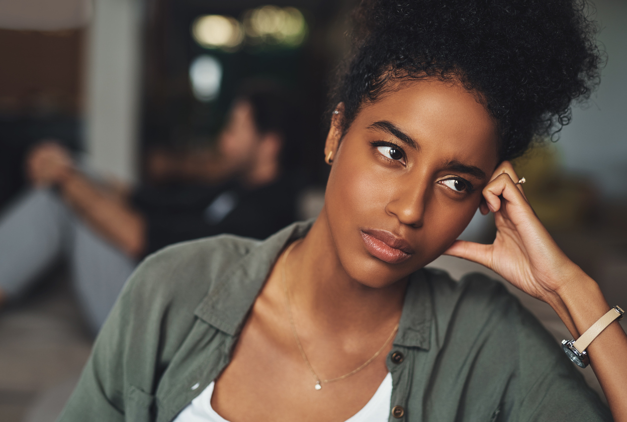 closeup of a woman resting her head on her hand looking stressed with her husband sitting quietly in the background