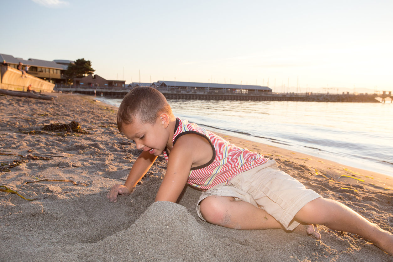 A young boy in shorts and a tank top plays in the sand on the beach in Edmonds, Washington. It's a sunny evening and a pier is visible behind him.