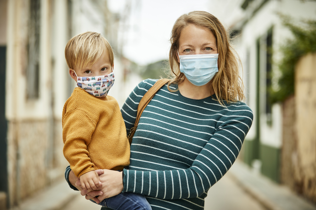 mother holding her son outdoors, both of them smiling under masks