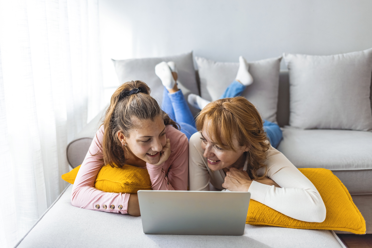 A mom and daughter lay on the couch on their bellies and look at a laptop; they're playing a mental health conversation starter game