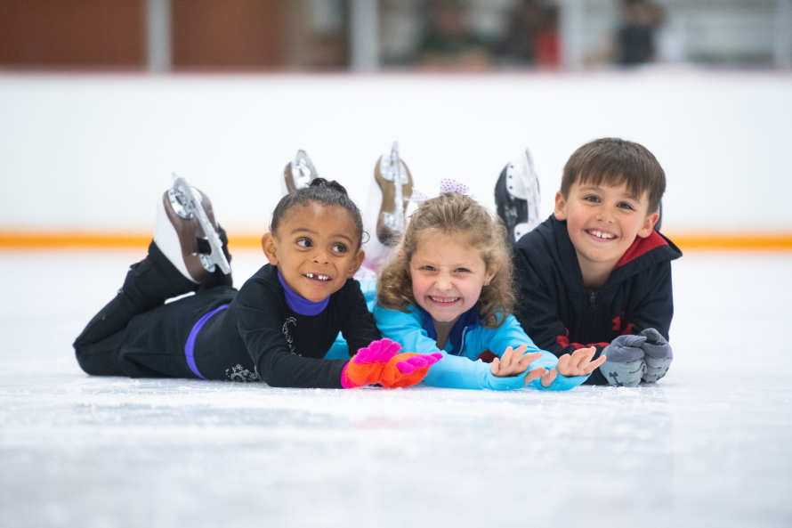 Three kids wearing ice skates lying on the ice smiling at the camera