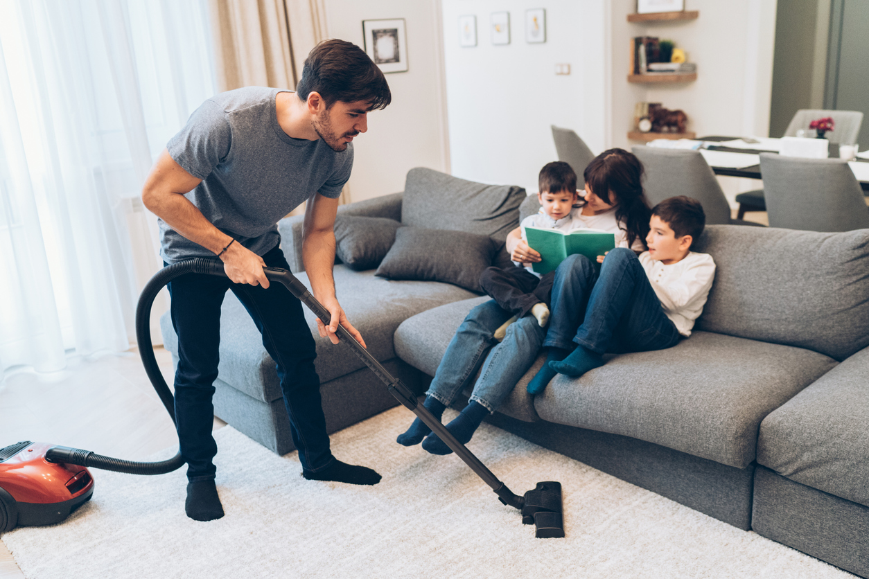 Father vacuums while mother reads a book to their kids