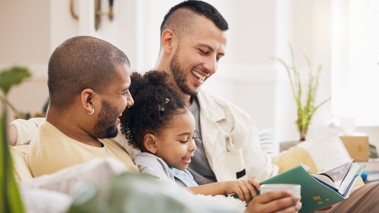 Two dads sitting on the couch with their daughter reading a book with LGBTQ characters