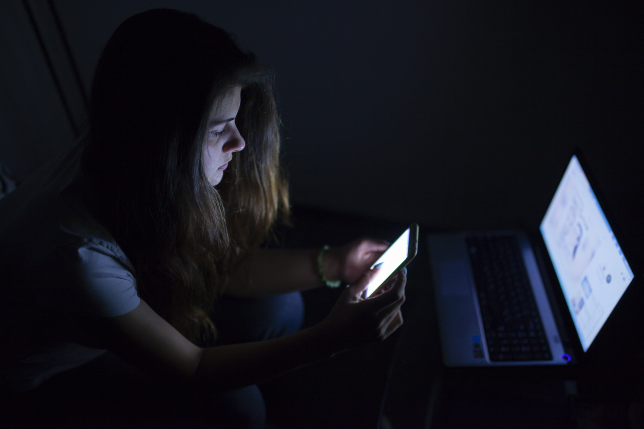 teenage girl in the dark with her laptop and phone screens lit up