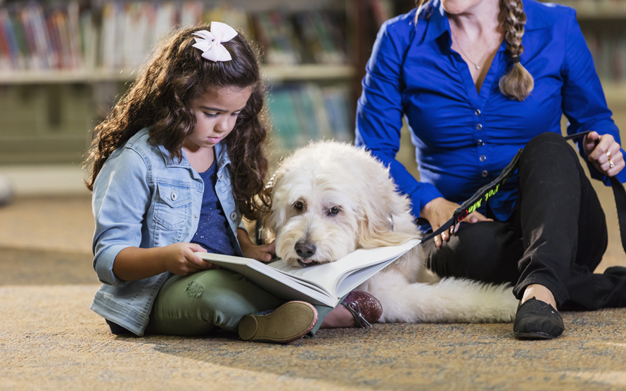 little girl reading to a dog in the library with a librarian nearby