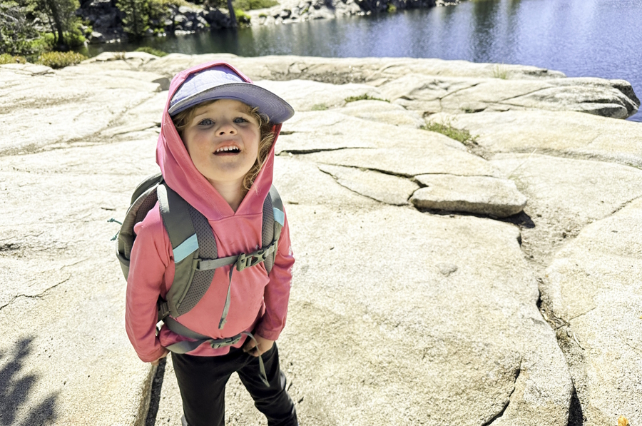 Young girl wearing a pink sweatshirt and a hat on a hike near a lake smiling for the camera