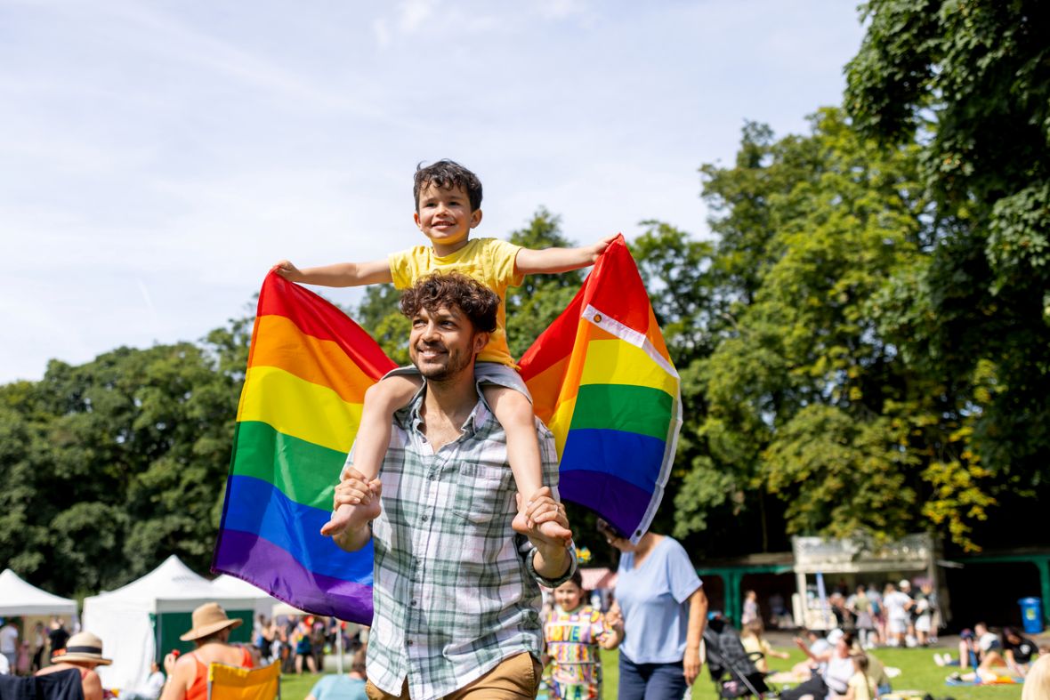 A young boy holds a Pride rainbow flag while on his father's shoulders to celebrate Pride.