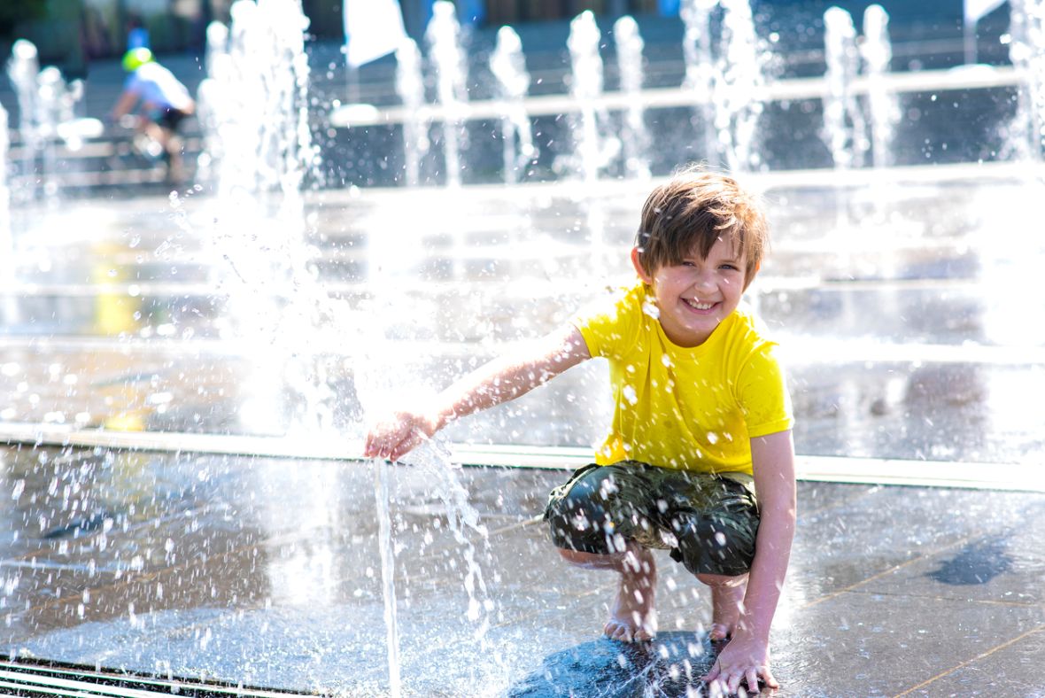 A young boy in a yellow shirt crouches down at a splash pad and enjoys the water.