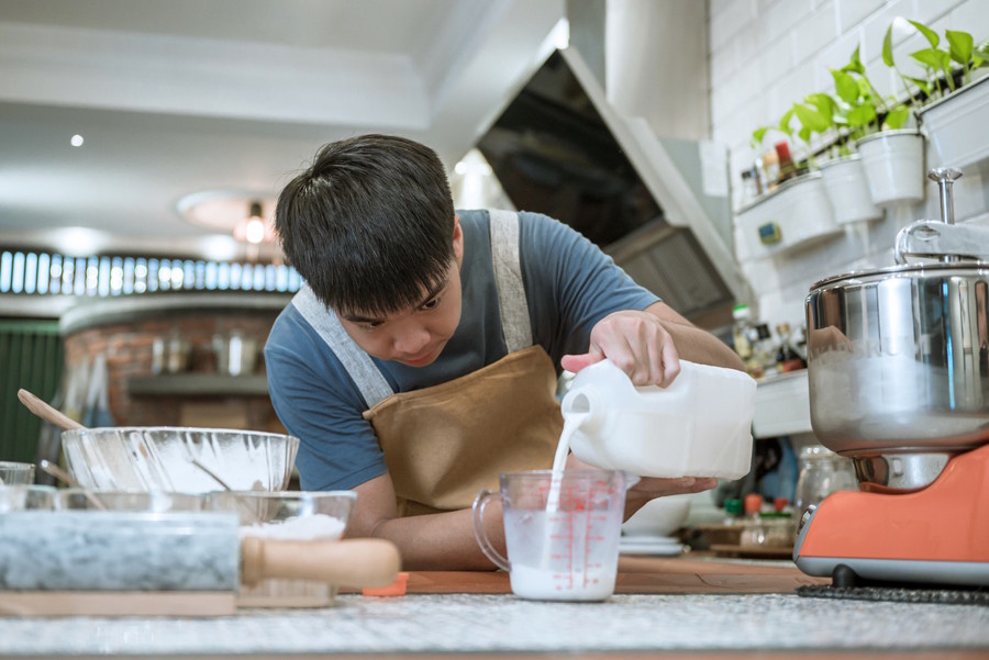 A teen boy is pouring milk into a measuring cup while baking in a kitchen