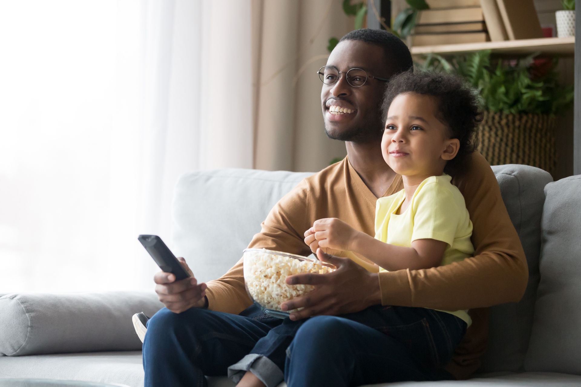 Happy black single father and cute little son sit on couch relax watching TV eating popcorn together, smiling african American dad and preschooler child enjoy family weekend with snack and cartoon