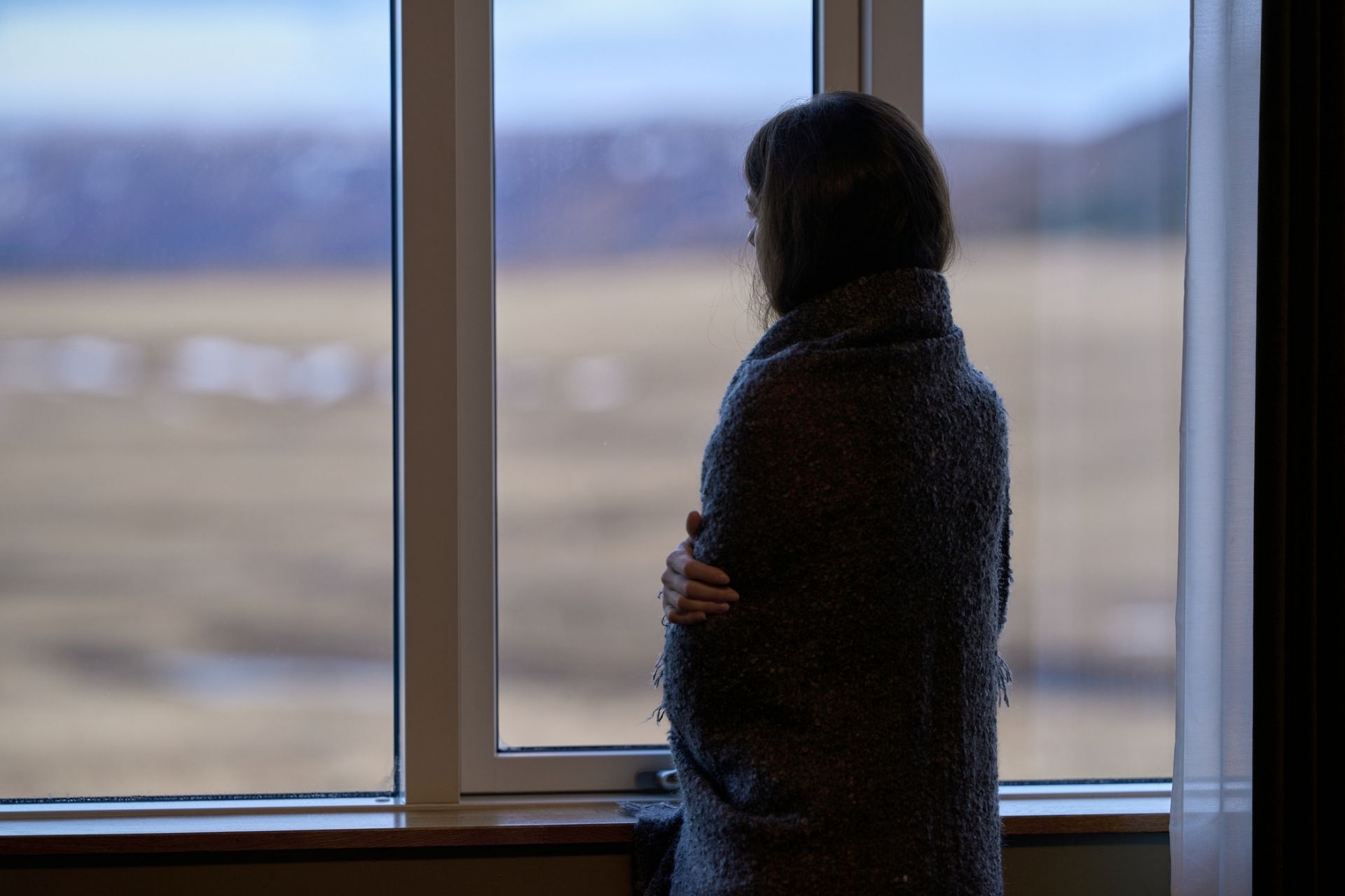 Woman wrapped in blanket looking out window at overcast landscape